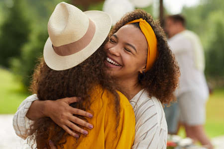 Portrait of young African-American woman hugging friend and smiling cheerfully while enjoying outdoor party in Summerの写真素材
