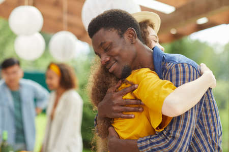 Waist up portrait of young African-American man hugging friend fondly while greeting each other at outdoors party in Summer, copy spaceの写真素材