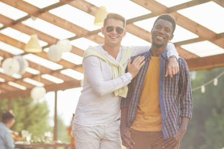 Waist up portrait of young men smiling at camera in sunlight, while posing outdoors in Summer with friends and family background, copy spaceの写真素材