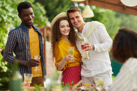 Portrait of young couple embracing while standing by table and enjoying dinner with friends outdoors...