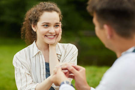 Close up portrait of beautiful young woman looking at boyfriend while putting on engagement ring on...