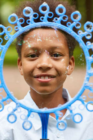 Vertical close up portrait of smiling African-American boy looking at camera through big bubble wand...