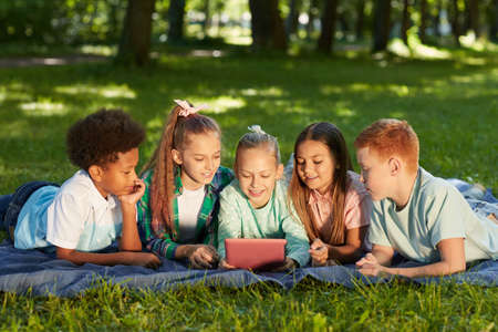 Front view at multi-ethnic group of kids using digital tablet while lying on green grass in park outdoors lit by sunlight, copy spaceの写真素材