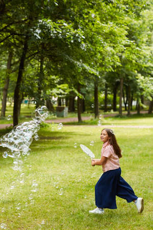 Vertical full length portrait of carefree girl running in park outdoors while playing with bubbles...