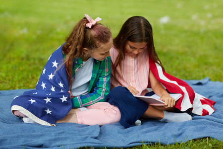 Full length portrait of two cute girls covered by American flag sitting on picnic blanket in park...