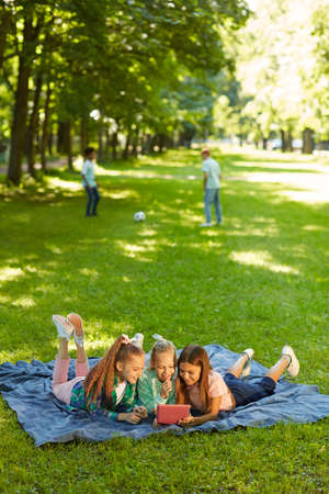 Vertical full length portrait of three teenage girls using digital tablet while lying on green grass in park outdoors lit by sunlight, copy spaceの写真素材