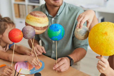 Close up of male teacher holding planet models and looking at camera while working with group of...