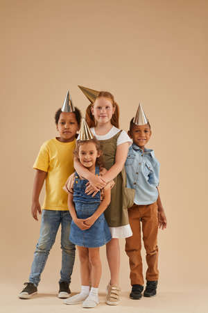 Vertical full length portrait of diverse group of children wearing party hats looking at camera while standing against plain background in studioの写真素材