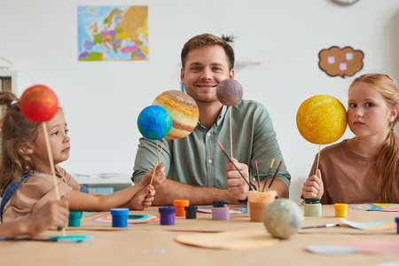 Portrait of smiling male teacher holding planet models and looking at camera while working with...