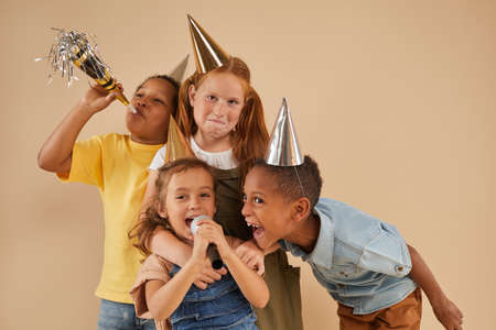 Waist up portrait of diverse group of children wearing party hats screaming to microphone while standing against plain background in studioの写真素材