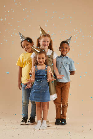 Vertical full length portrait of diverse group of children wearing party hats smiling at camera...