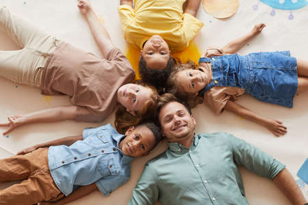 Above view at smiling male teacher with multi-ethnic group of children lying in circle and looking...