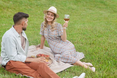 High angle portrait of beautiful blonde woman holding wineglass while enjoying picnic on green grass...