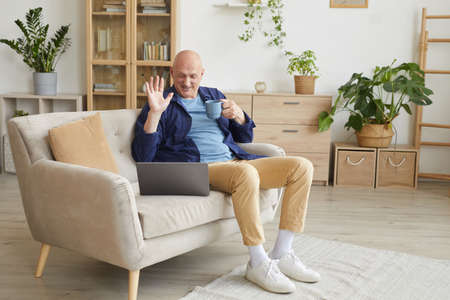 Full length portrait of modern senior man waving at camera and smiling happily during video call at...