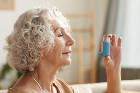 Side view close up portrait of senior woman using inhaler for asthma or breathing problems in home settingの写真素材