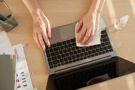 Above view close up pf unrecognizable woman sanitizing laptop while working at desk in post pandemic...