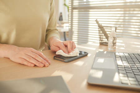 Close up pf unrecognizable woman sanitizing smartphone while working at desk in post pandemic office...