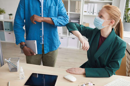 Portrait of young businesswoman wearing mask bumping elbows with coworker as contactless greeting in...
