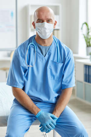 Vertical medium long portriat of unrecognizable doctor wearing blue uniform, latex gloves and mask on face sitting relaxed looking at cameraの写真素材