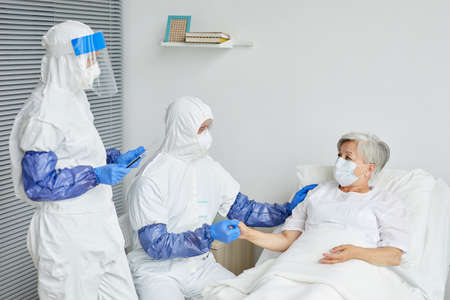 Two professional doctors wearing protective suits with respiratory masks and gloves examining senior...