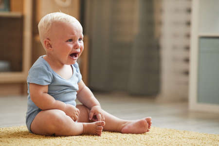 Horizontal shot of sad little baby boy sitting on floor in living room crying, copy spaceの写真素材