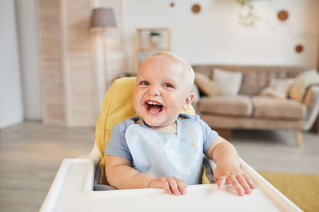 Horizontal portrait of cheerful little child sitting on high chair with food around his mouth...