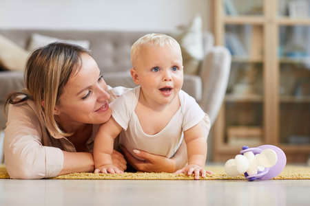 Happy young mother spending time at home lying on carpet with her little baby son, horizontal shot