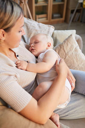 Vertical high angle shot of modern Caucasian woman sitting on sofa cuddling her lovely baby son in...