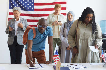 Multi-ethnic group of people registering at polling station decorated with American flags on...