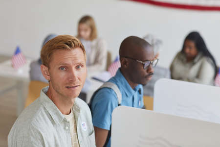 High angle portrait of modern blonde man looking at camera while standing in voting booth on...