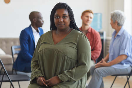 Portrait of young African-American woman looking at camera during support group meeting with people...