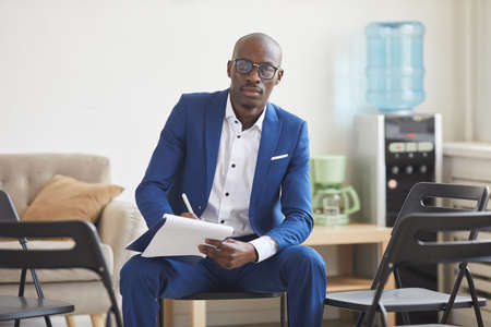 Portrait of elegant African-American man holding clipboard and looking at camera while sitting on...