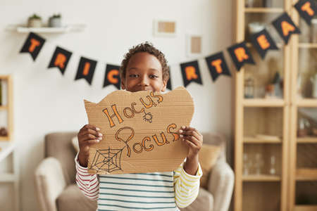 Modern joyful African American boy standing in living room decorated for Halloween holding cardboard with Hocus Pocus letteringの写真素材