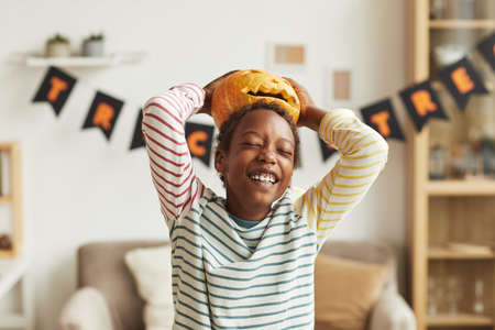 Playful African American boy having fun with Jack O Lantern pumpkin putting it on his head, medium portrait shotの写真素材