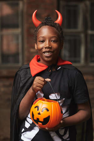 Vertical medium portrait of happy African American boy dressed up as devil with red horns holding basket full of candies after trick-or-treatingの写真素材