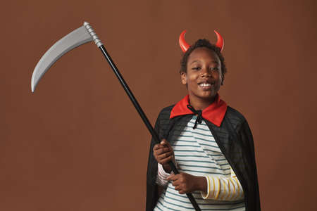 Studio portrait of cheerful African American preteen boy wearing red horns on head and costume mantle holding scythe looking at camera smiling, brown backgroundの写真素材