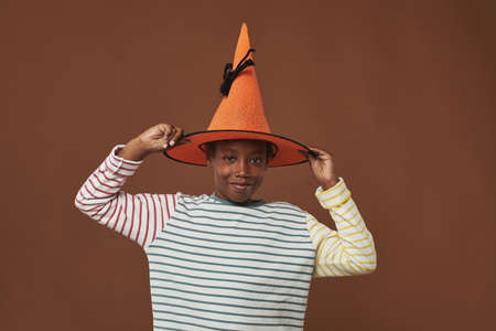 Studio portrait of cheerful young boy standing against brown wall background trying on funny orange...