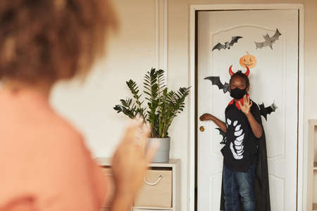 Over-the-shoulder shot of young mother seeing off her little son who is wearing devil Halloween costume going out for trick-or-treating with friends.の写真素材