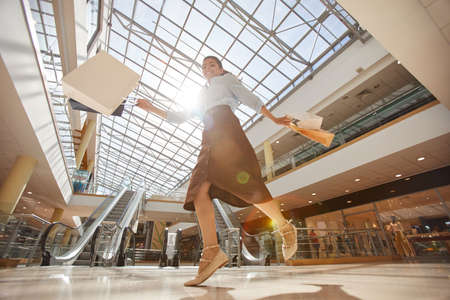 Warm-toned low angle portrait of happy young woman jumping in mall and holding shopping bags, copy spaceの写真素材
