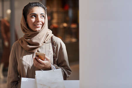 Waist up portrait of beautiful Middle-Eastern woman looking away lit by sunlight while enjoying shopping in mall, copy spaceの写真素材