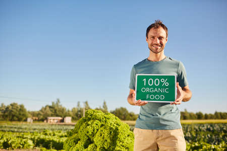 Portrait of smiling man holding ORGANIC FOOD sign and looking at camera while standing at vegetable plantation outdoors in sunlight, copy spaceの写真素材