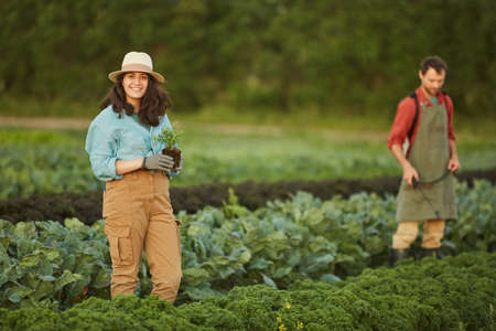 Portrait of two people working in field at vegetable plantation, focus on young woman smiling at camera in foreground, copy spaceの写真素材