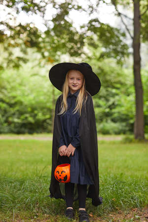 Vertical full length portrait of smiling teenage girl dressed as witch posing outdoors and holding Halloween bucketの写真素材