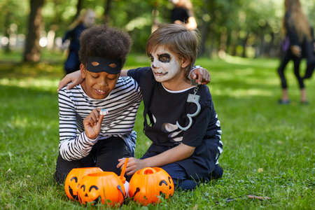 Full length portrait of two boys wearing Halloween costumes sitting on green grass outdoors with candy buckets, copy spaceの写真素材