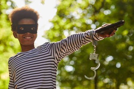 Waist up portrait of smiling African-American boy wearing bandit costume for Halloween and holding toy gun while posing outdoors, copy spaceの写真素材