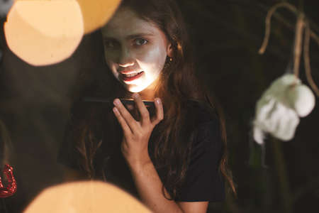 Portrait of smiling teenage girl looking at camera and holding flashlight during outdoor Halloween...