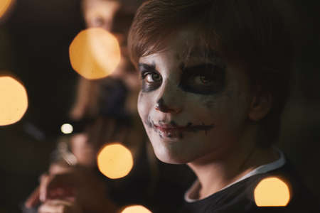 Portrait of teenage boy wearing face paint and looking at camera during outdoor Halloween party with...