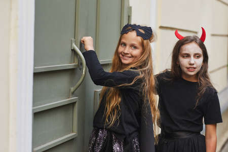 Portrait of two girls wearing Halloween costumes knocking on doors and smiling at camera while trick...