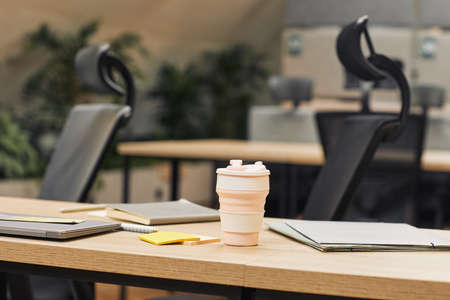 Close up background image of modern open space office decorated with plants, focus on coffee cup on...