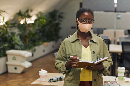 Waist up portrait of young African-American woman wearing mask and looking at camera while standing in post pandemic office, copy spaceの写真素材